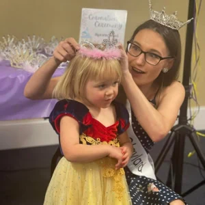 A girl wearing a crown is placing a crown on a little girl's head.